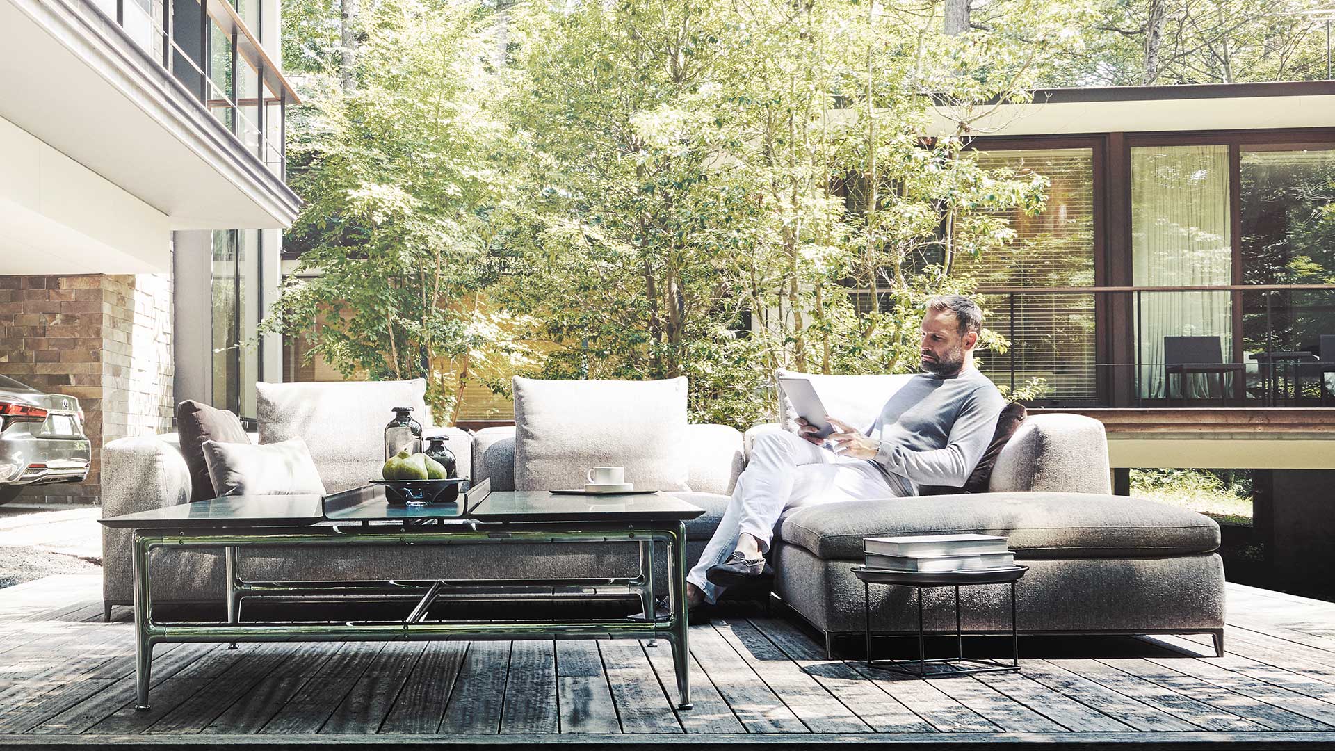 Man seated in an outdoor couch while reading in front on a luxury home