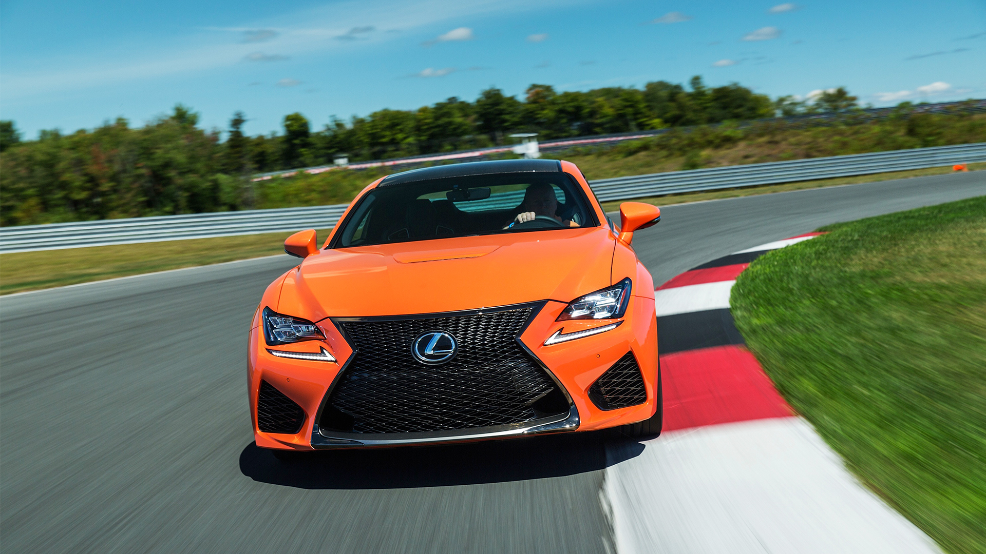 An orange RC F coupe driving around a race track with blue sky and green trees in the background